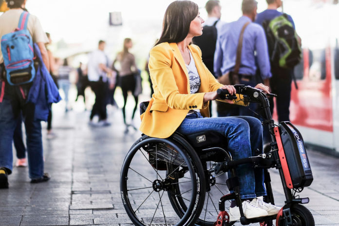 Frau sitzt im Rollstuhl mit elektrischem Zuggerät davor am Bahnsteig. 