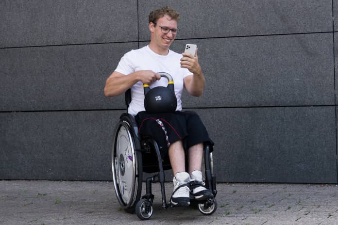 Auf dem farbigen Foto ist ein Mann im sportlichem Outfit im Rollstuhl sitzend zu sehen. Er schaut auf sein Handy, welches er in der linken Hand hält. In der anderen hält er eine Hantel (Kettlebell) auf seinem Schoß fest.