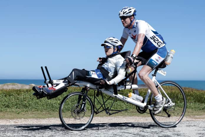 Auf einer Art Cargo-Rad fahren Vater und Sohn eine Straße entlang, beide in Wettkampfkleidung. Der Sohn ist körperlich behindert, sitzt vorne, hat jedoch keine Pedale zum treten.