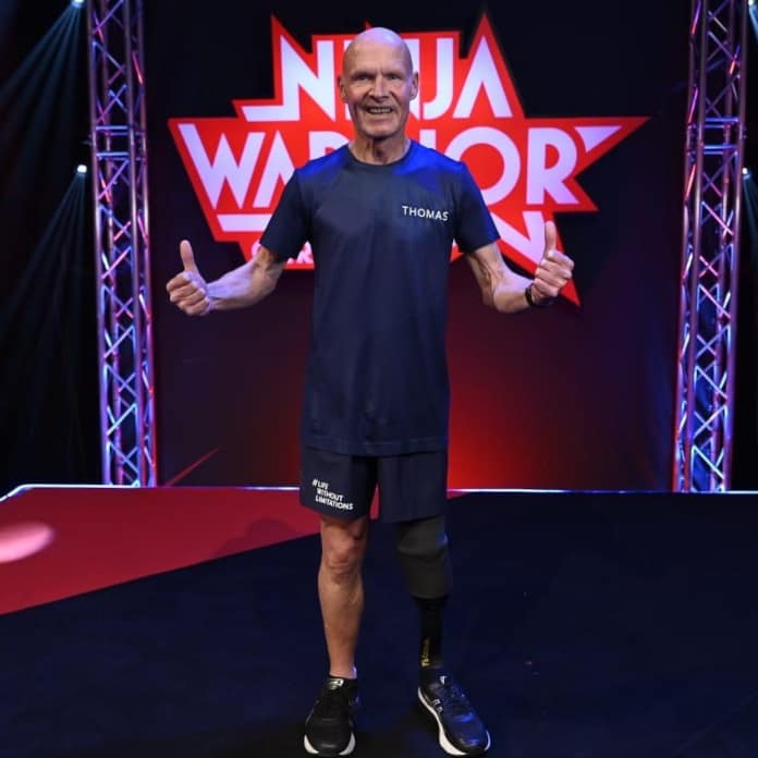 Thomas Wedig steht vor dem Ninja-Warrior-Logo bei der Show und hält beide Daumen in die Höhe.