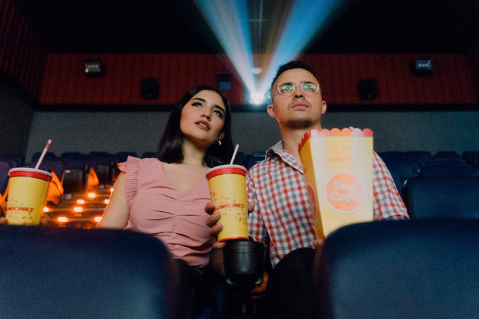 Eine Frau und ein Mann sitzen auf Kinosesseln in einem Saal und schauen gespannt nach vorn auf die Leinwand. Der Mann hält eine Popcorntüte in der rechten Hand und ein Getränk