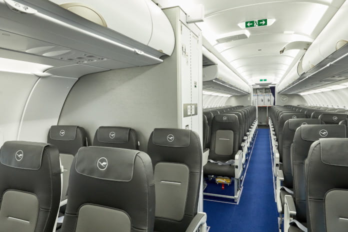 Gang an Bord eines Flugzeuges, mit den Sitzen links und rechts. Auf der linken Seite ist nach der 2. Sitzreihe das Bord-WC zu finden. Zu sehen ist die Tür.