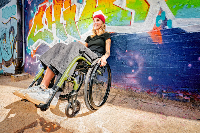 Eine junge Frau lehnt mit ihrem Rollstuhl an einer Wand voller Graffiti. 