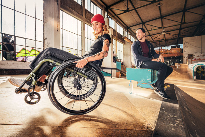 Eine junge Frau sitzt in dem Rollstuhl Nano X, den sie leicht kippt. Im Hintergrund ist eine Industriehalle zu sehen. Neben ihr sitzt ein Mann auf einem Holzblock.