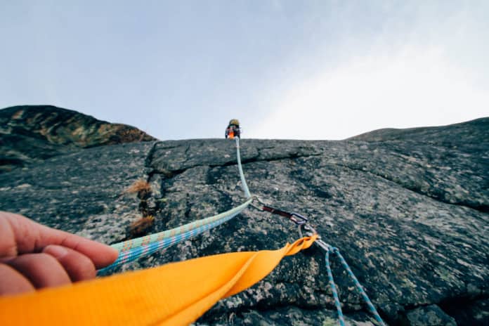 Eine Hand hält ein Seil an einer Felswand fest, daneben ist ein zweites Seil zu sehen. Weiter oben an der Felswand klettert ebenfalls ein Mensch.