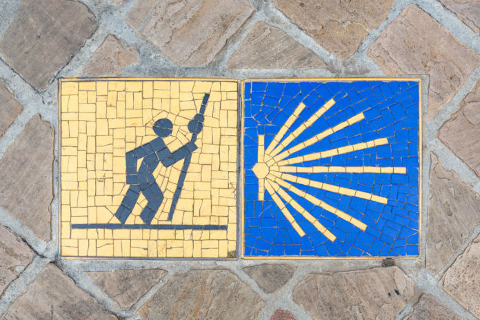 Mosaikbild auf einem Boden in zwei farbige Quadrate unterteilt. Erstes Quadrat: Ein Männchen mit Pilgerstock auf gelben Hintergrund. Zweites Quadrat: Gelbe Jakobsmuschel auf blauen Untergrund.