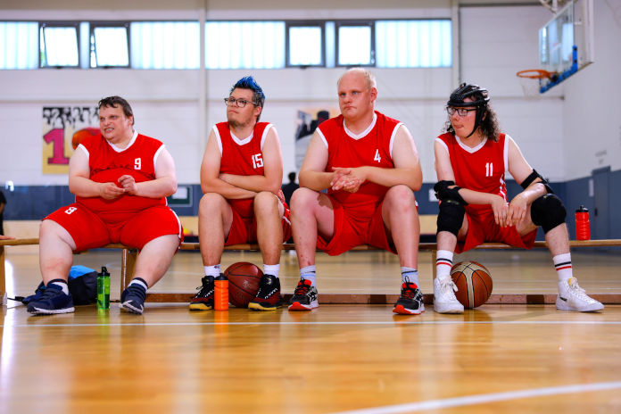 4 Männer mit sichtbarer Behinderung sitzen in Sporttrikots auf einer Bank in einer Sporthalle und schauen in eine Richtung. 