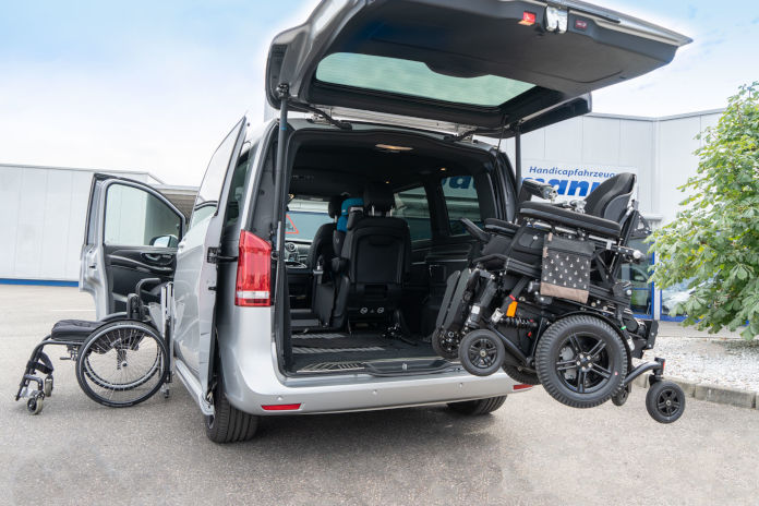 Eine silberne Mercedes V-Klasse ist mit 2 Verladesystemen für einen Rollstuhl ausgestattet: Einmal für die seitliche Verstauung sowie mit der Möglichkeit, einen Rollstuhl mit dem Kran in den Kofferraum verladen zu können.