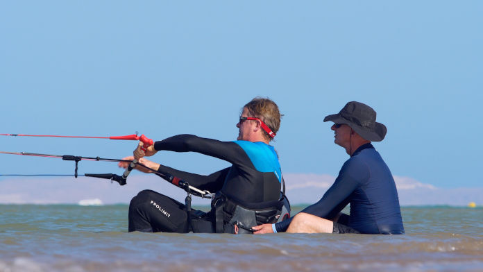 Zwei Männer sitzend hintereinander beim Kitetraining im flachen Wasser. Der vordere Mann hält sich an einer Leine fest. Der hintere gibt Hilfestellung und hält ihn am Trapez (Hüftgürtel) fest. 