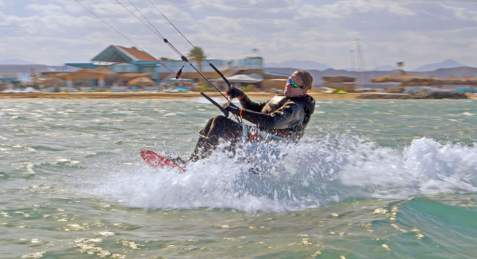 Ein Mann hält sich an einer Leine fest und wird sitzend auf einem Kiteboard rasant durchs Wasser gezogen. Das Wasser spritzt.  