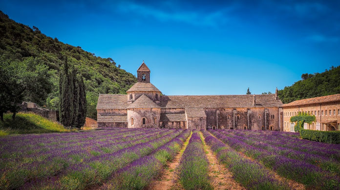 Das Bild zeigt ein altes Kloster inklusive einem Anbau, umgeben von Lavendelreihen im Vordergrund des Bildes. Blauer Himmel und Bäume an der rechten und linken Seite des Bildes lassen es an die Toscana erinnern.