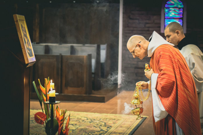 Das Foto zeigt einen Mönch leicht gebeugt vor einem Bild stehend. In der Hand hält er ein Schwenkrauchgefäß. Der Rauch steigt aus dem Gefäß empor. Neben ihm steht ein weiterer Mönch.