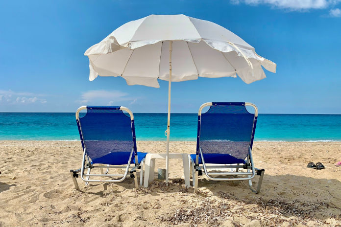 Zwei Sonnenstühle stehen am Strand, davor ein Sonnenschirm. Im Hintergrund das Meer.