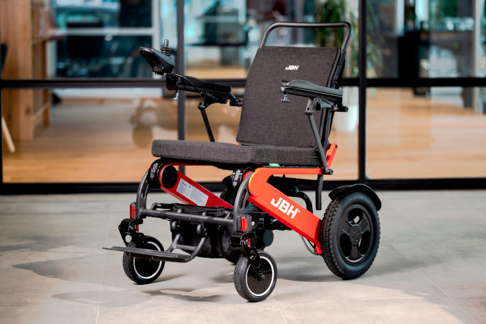 Das Foto zeigt einen faltbaren E-Rollstuhl mit breiter Sitzfläche. Teile des Untergestells sind orange/rotfarben. Die beiden hinteren Räder sind deutlich größer als die vorderen. An der rechten Armlehne befindet sich ein Joystick.