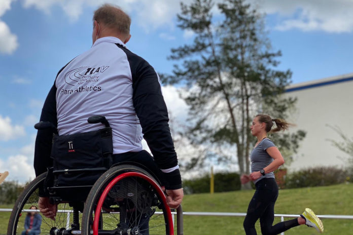 Ein Rollstuhlfahrer, der an einer Laufbahn steht und eine vorbei laufende Frau beobachtet. Auf seiner Trainingsjacke steht das Wort Para-Athletics.