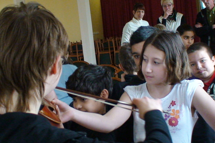 ein Mädchen berührt mit ihrer Hand eine Geige von einer Musikerin, im Hintergrund viele Menschen