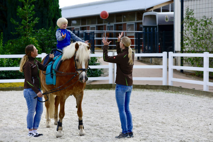 Das Foto zeigt ein Kind auf einem Pferd sitzend. Eine Therapeutin steht vor dem Pferd und wirft dem Kind einen Ball zu. Links neben dem Pferd steht eine Frau und hält das Pferd an den Zügeln fest. Sie befinden sich auf einem Reitplatz.