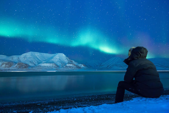Eine Person am Meeresufer sitzend. Am Himmel Polarlichter in grün und Blautönen. Darunter schneebedeckte Berge.