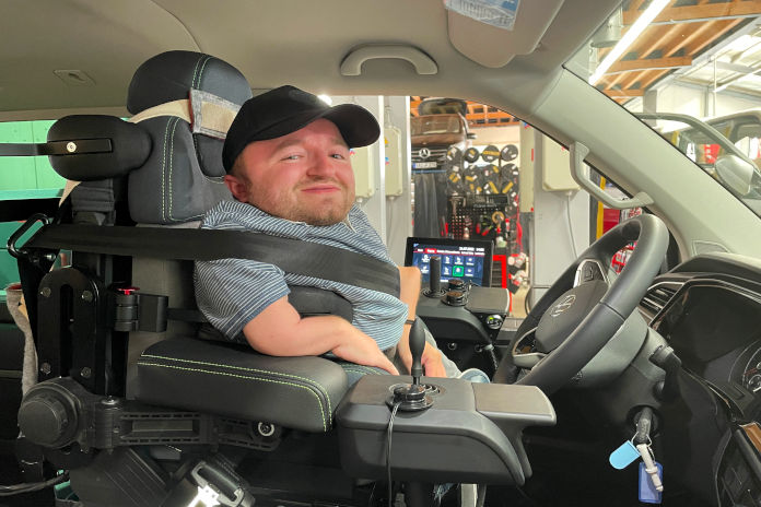 Ein Mann mit sichtbarer körperlicher Behinderung sitzt in einem Auto hinter dem Steuer. Neben seinem Arm ist eine Konsole mit einem Joystick sichtbar.