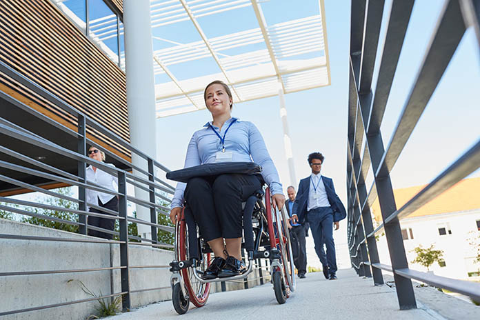 Eine junge Geschäftsfrau im Rollstuhl zwischen zwei Geländern fahrend. Auf ihren Oberschenkeln liegt eine Laptoptasche. Im Hintergrund laufen weitere Personen.