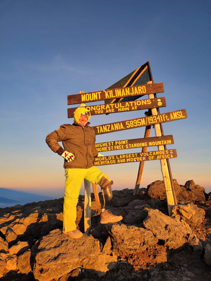 Das Bild zeigt einen Mann stehend, angelehnt an ein Schild auf dem Gipfel des Kilimandscharo. Links trägt er eine sichtbare Beinprothese.