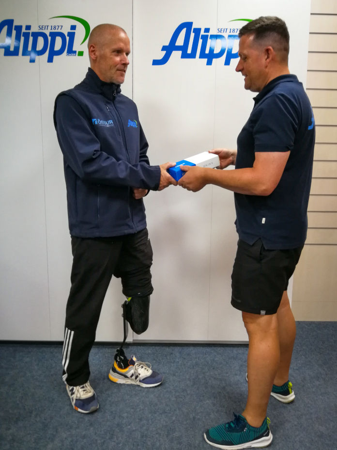 Das Bild zeigt Jens König und Techniker Thomas Schröder gegenüberstehend. Jens König trägt eine Unterschenkelprothese am linken Bein. Eine Schachtel wird übergeben.
