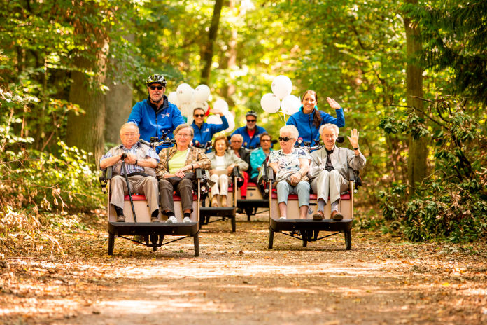 Das Foto zeigt eine fröhliche Runde älterer Menschen, die in einem Triobike sitzen. Sie werden durch die Fahrer:innen durch ein Waldstück gefahren.