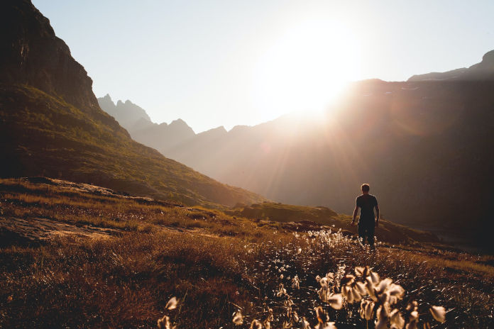 Eine Person läuft bei untergehender Sonne an einem Bergfuß entlang über eine Wiese.