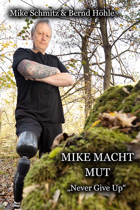 Ein Buchcover mit dem Titel Mike macht Mut Never give up. Ein Mann steht mit den Armen verschränkt in einem Waldstück. Er trägt am rechten Bein eine Prothese.
