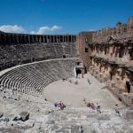Ein rundes, antikes Amphitheater. Ganz oben Torbögen darunter Sitzreihen im Vordergrund eine Bühne. Im inneren Verzierungen aus Ornamenten und Säulen.