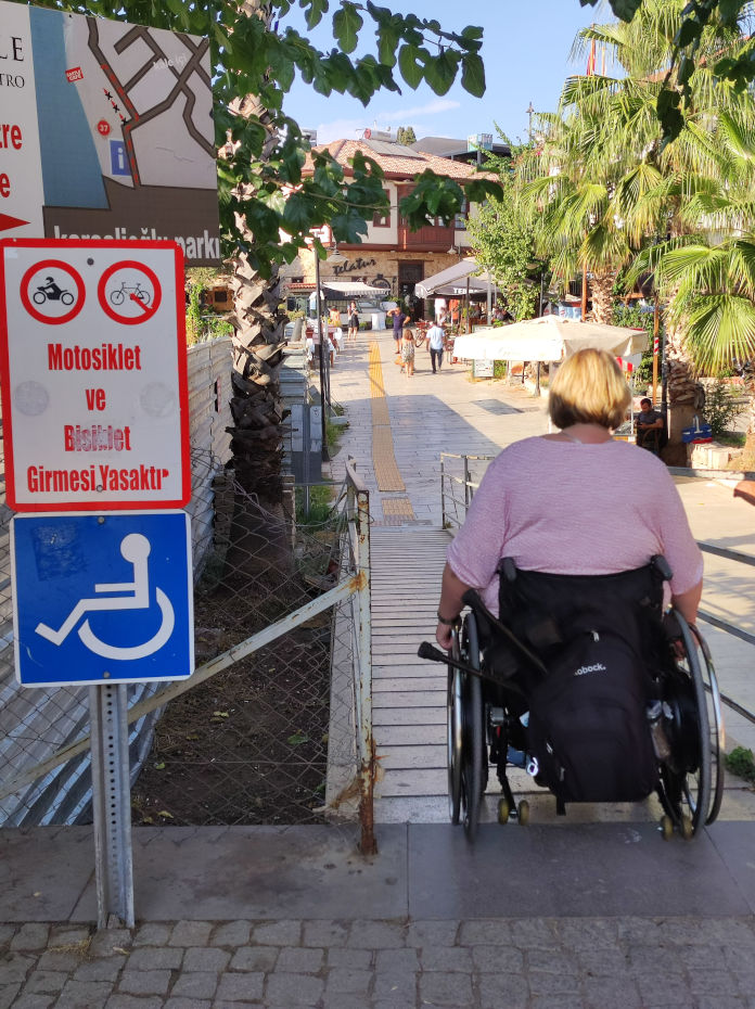 Eine Rollstuhlfahrerin die eine Rampe mit Geländer herunterfährt. Ein blaues Schild mit weißem Rollstuhl und ein Warnschild das die Benutzung der Rampe für Fahrräder und Motorräder untersagt. 