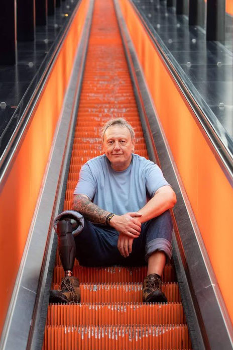 Ein Mann sitzt auf einer Stufe einer Rolltreppe. Er trägt an seinem rechten Bein eine Prothese. Die Stufen der Rolltreppe sind orangefarben.