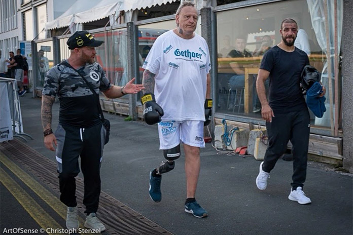 Drei nebeneinander laufende Männer. Der Mann in der Mitte trägt am rechten Bein eine Prothese und angezogene Boxhandschuhe. Die Männer neben ihm sind sportlich gekleidet.