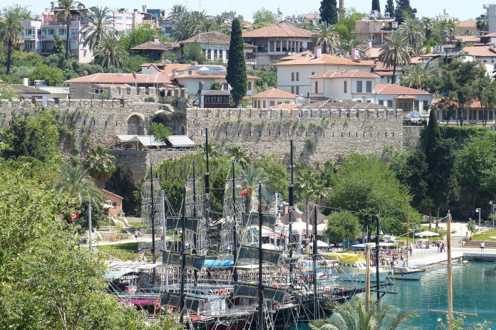 Ein Hafen mit Schiffen. Weiter oben eine Steinmauer umgeben von Palmen und Wohnhäusern.