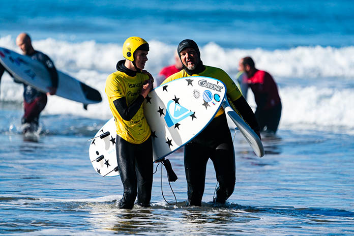 Zwei Männer nebeneinander im flachen Wasser laufend. Sie tragen ihre Surfbretter unterm Arm. Im Hintergrund weitere Surfer und Wellen.