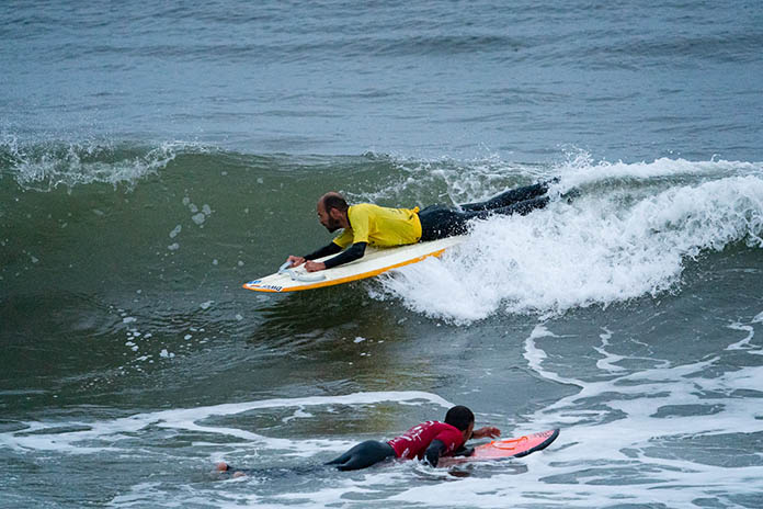 Zwei Männer liegend auf Surfbrettern entgegengesetzt surfend. Der vordere Mann in die Welle hinein. Der hintere Mann mit der Welle. 