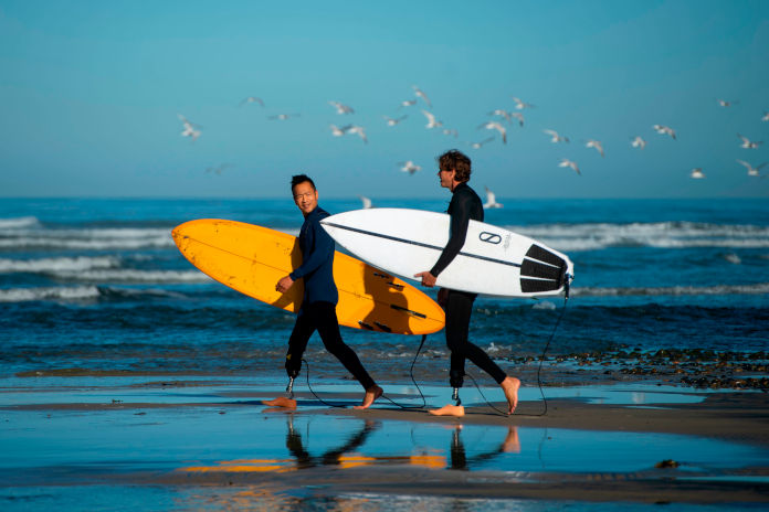 Zwei männliche Surfer an einem Strand laufend. Beide tragen ein Surfbrett unter dem Arm. Beide tragen jeweils eine Beinprothese.