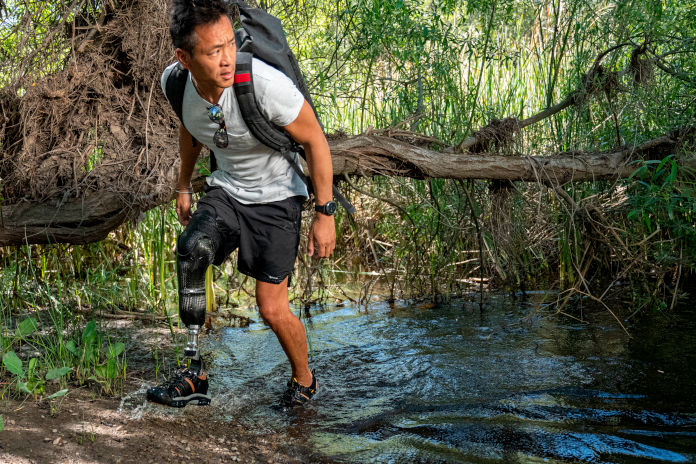 Ein Mann läuft aus einem Fluss heraus. An seinem rechten Bein trägt er eine Prothese. Sein linker Fuss steht noch im Wasser. Im Hintergrund Buschwerk und Bäume.