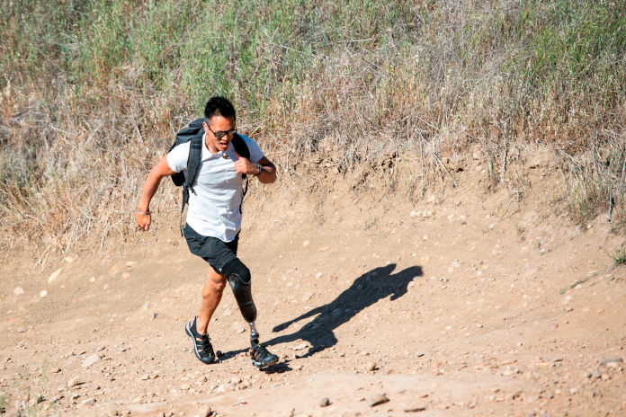 Ein Mann läuft einen steinigen Sandweg in einer Kurve hinauf. Er trägt am linken Bein eine Prothese. Auf dem Rücken trägt er einen Rucksack.