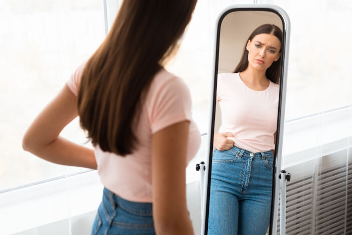 Eine Frau schaut sorgenvoll in den Spiegel und betrachtet ihren Körper.