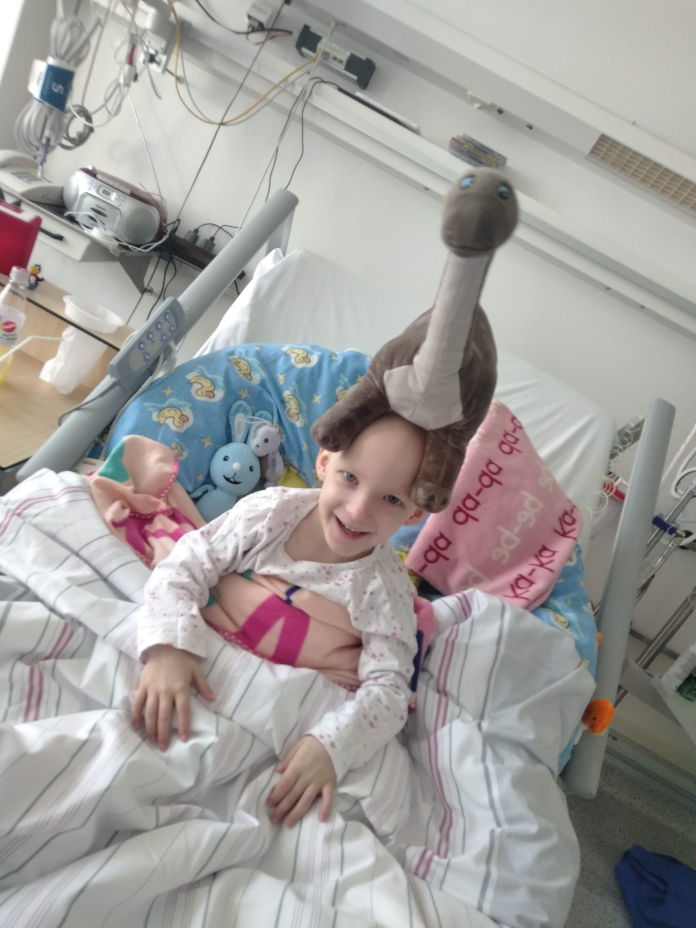 Ein kleines Mädchen sitzt in einem Krankenhausbett, auf dem Kopf ein Plüschdinosaurier. 