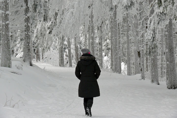 Ein Winterbild. Eine Frau läuft im Wald durch Schnee. Sie trägt einen schwarzen Wintermantel, schwarze Winterstiefel und eine graue Mütze. Umgeben ist sie von schneebedeckten Bäumen.