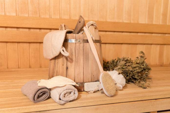 Eine Sitzfläche in einer Sauna. Darauf steht ein Aufgusseimer, liegen zusammengerollte Handtücher, ein Birkenquast und verschiedene Wellnessartikel.