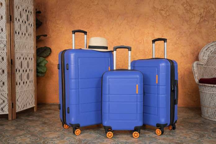 Drei blaue Koffer in unterschiedlichen Größen und orangefarbenen Rollen stehen in einem Raum. Auf einem Koffer liegt ein Sonnenhut.