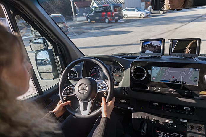 Eine junge Frau hinter einem Steuer im Auto sitzend. Vor ihr auf der Mittelkonsole ein Kamerasystem mit Monitor angebracht. Darunter ein Navigationsgerät.