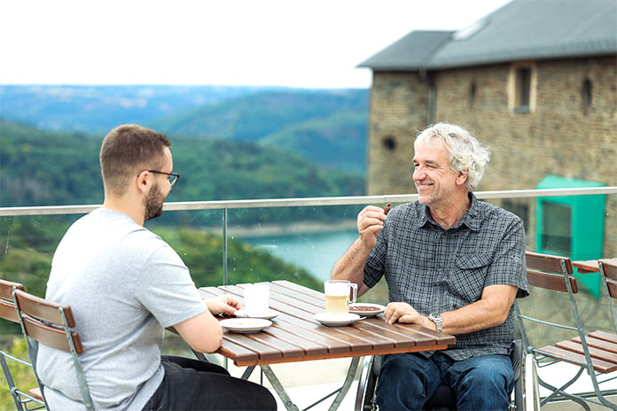 Zwei Männer sitzen an einem Tisch gegenüber voneinander. Ein Mann sitzt im Rollstuhl. Sie lächeln und trinken Kaffee. Der Ausblick geht über die Eifel hinweg.