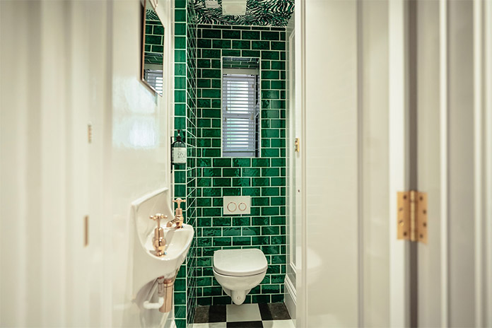 Ein kleines Badezimmer mit weißem WC, Waschbecken mit zwei goldenen Wasserhähnen. Hinter dem WC befinden sich grüne Fliesen und ein schmales Fenster.