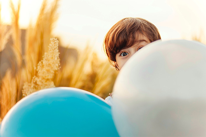 Ein kleiner Junge schaut mit seinem rechten, weit geöffneten braunen Auge über einen weißen Luftballon. Sein linkes Auge ist durch den Luftballon verdeckt. Er trägt braunes Haar. Links neben dem weißen Luftballon ist ein hellblauer Luftballon.