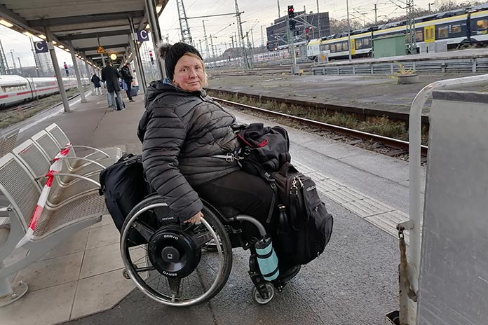 Am Bahngleis. Eine Frau sitzt im Rollstuhl. An dem Rückteil des Rollstuhls hängt ein Rucksack auf ihren Beinen liegt eine Tasche und an den Schienbeinen ist ein weiterer Rucksack befestigt.