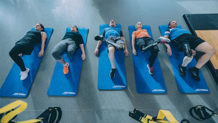 5 Menschen mit einer Amputation liegen auf blauen Sportmatten in einem Sportraum. Die Beine sind aufgestellt.